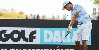 Golfer in light blue patterned shirt and white shorts taking a swing in front of a LIV Golf Dallas Championship sign